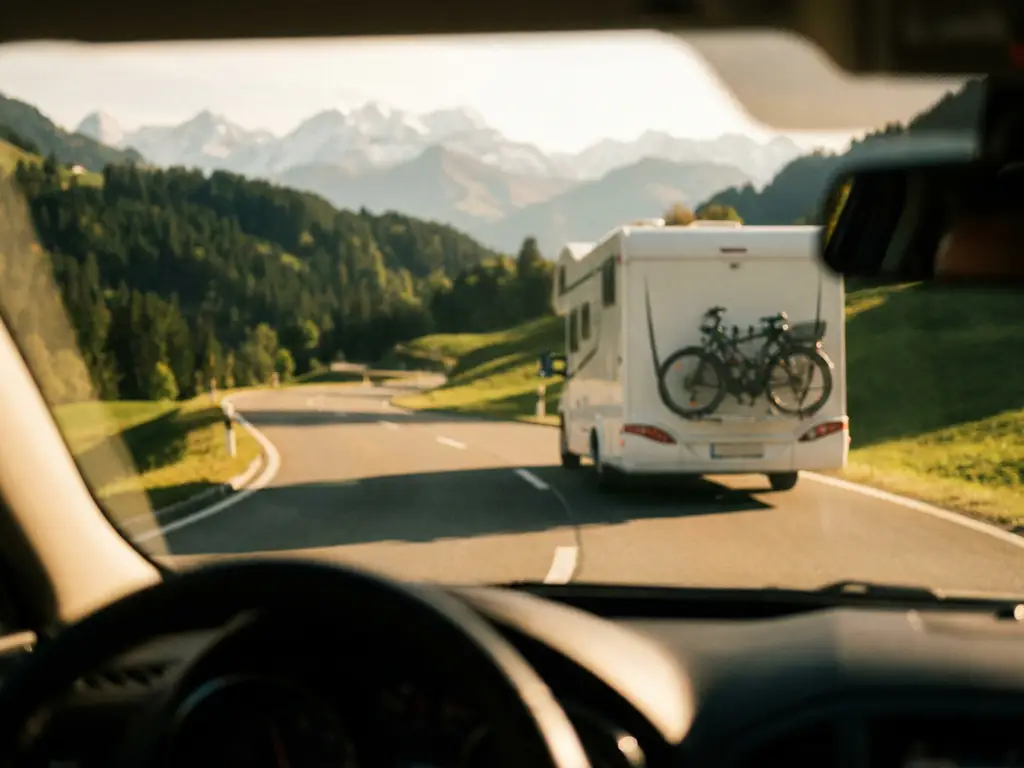 Witte camper op kronkelende Alpenweg met groene hellingen en bergtoppen op de achtergrond in middagzon.