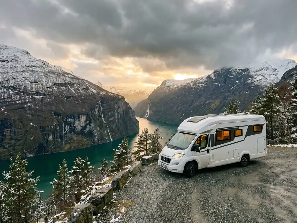 Witte campervan geparkeerd bij Noors fjord-uitkijkpunt, omringd door besneeuwd dennenbos en watervallen in gouden avondlicht.