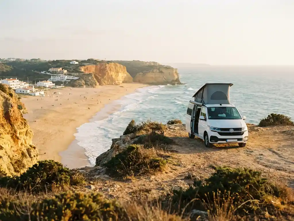 Witte campervan op rotsachtige kustlijn in Portugal met uitzicht op goudkleurig strand en turquoise Atlantische oceaan.