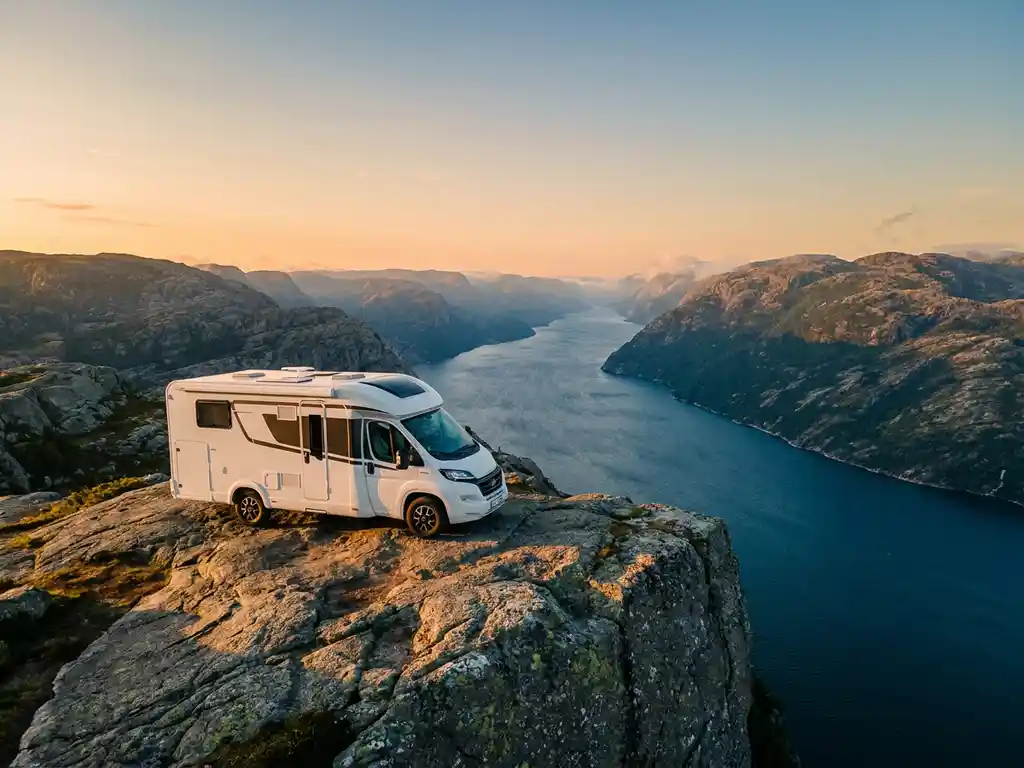 Witte campervan op rotsachtige Noorse kustklif met fjord en mistige bergen op de achtergrond tijdens gouden uur.
